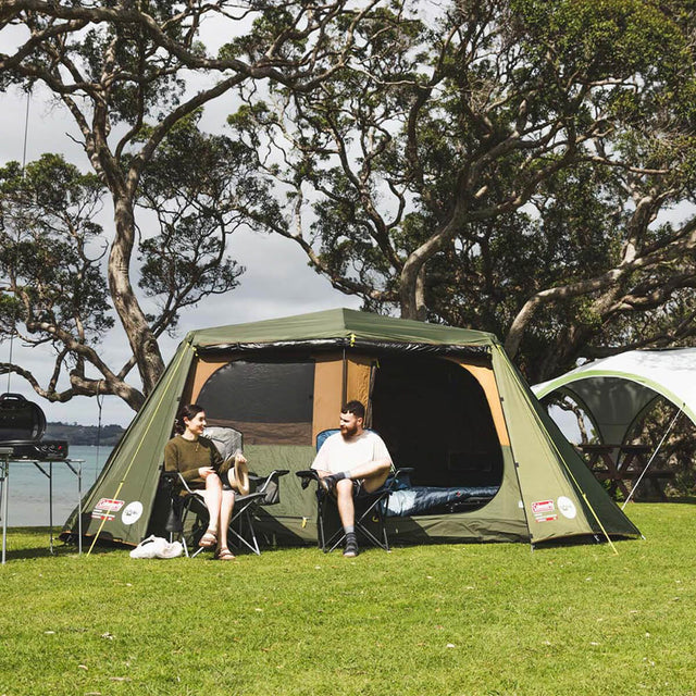 Front view of pitched tent set up near a beach with a couple in chairs outside, fly door rolled and one bedroom door open